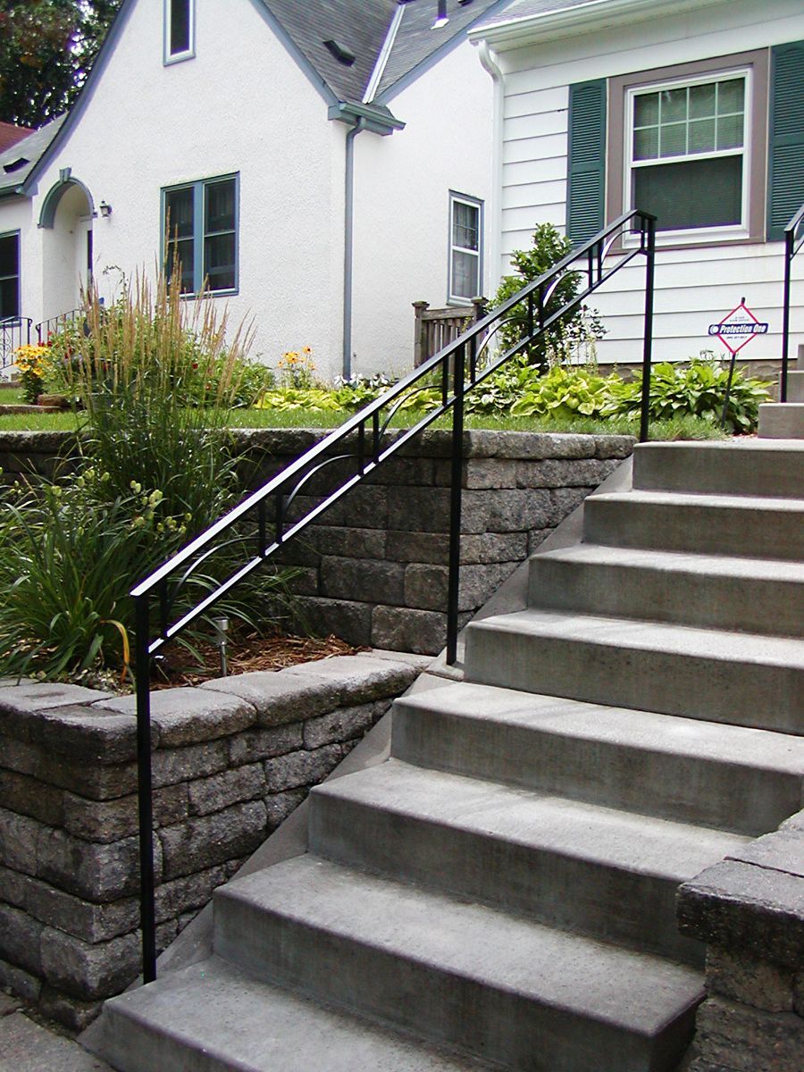 Black metal handrails on concrete steps leading to a white house with green shutters and stone retaining walls.