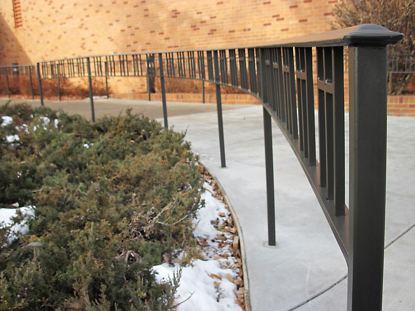 Black metal railing along a curved concrete path next to a hedge and brick building.