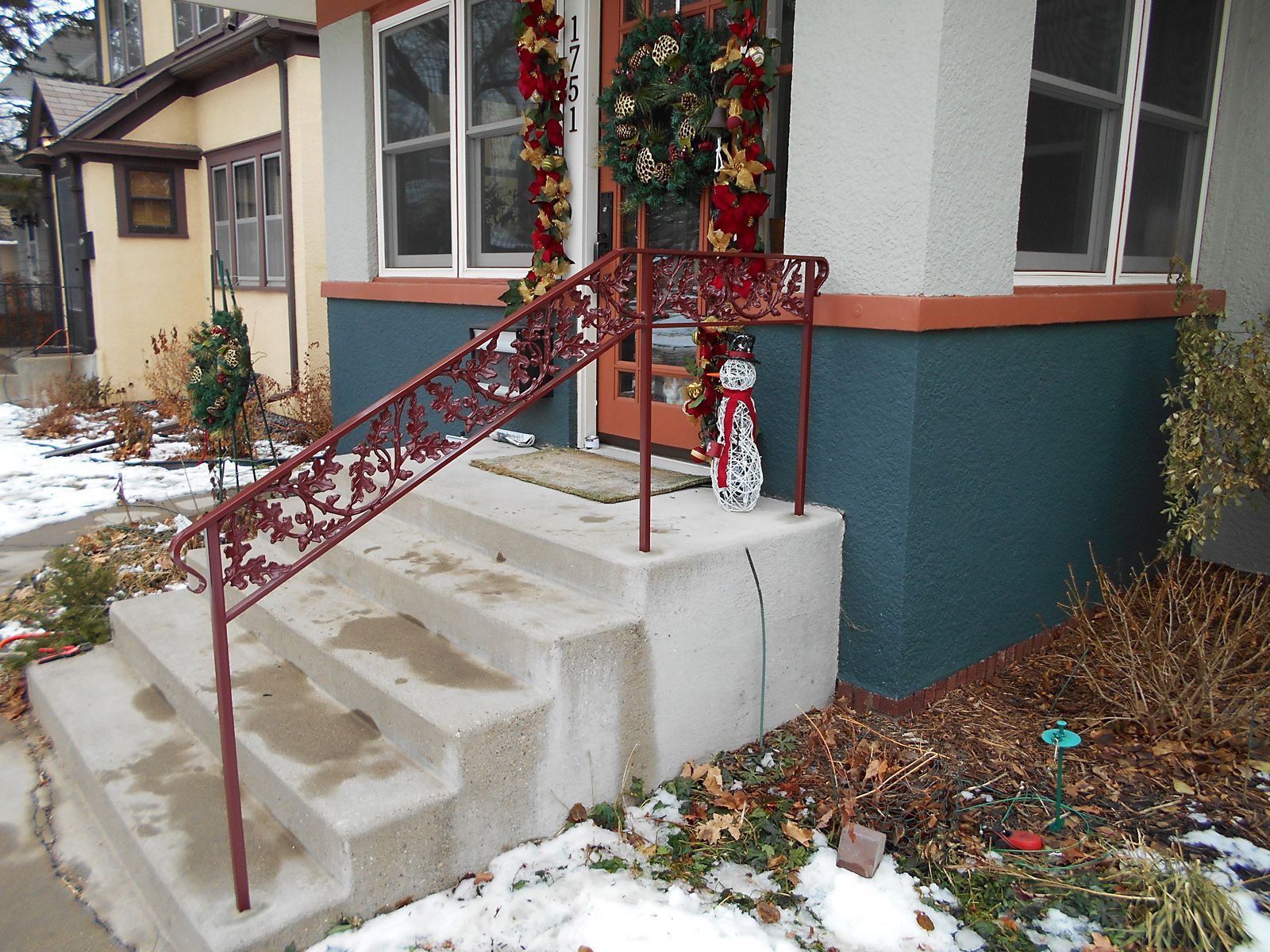 Red ornate railing on concrete steps leading to a decorated front door. Snow on the ground.