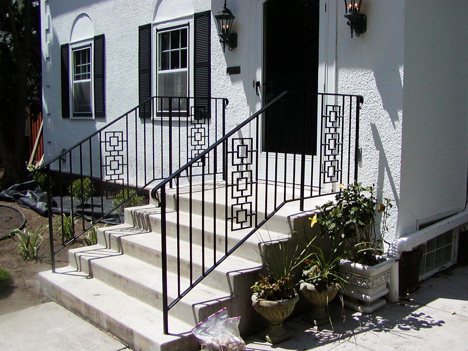 Black metal railing on concrete steps leading to a white stucco house with a black door.