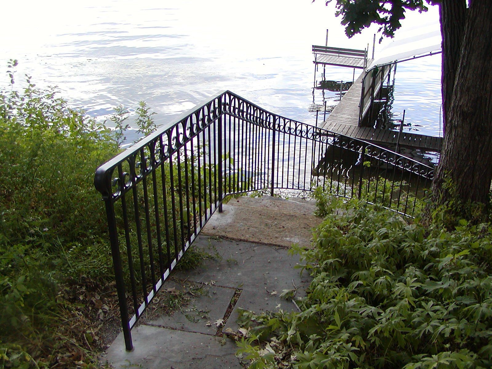 Staircase with black railing leading down to a dock on a lake. Overcast, green foliage.