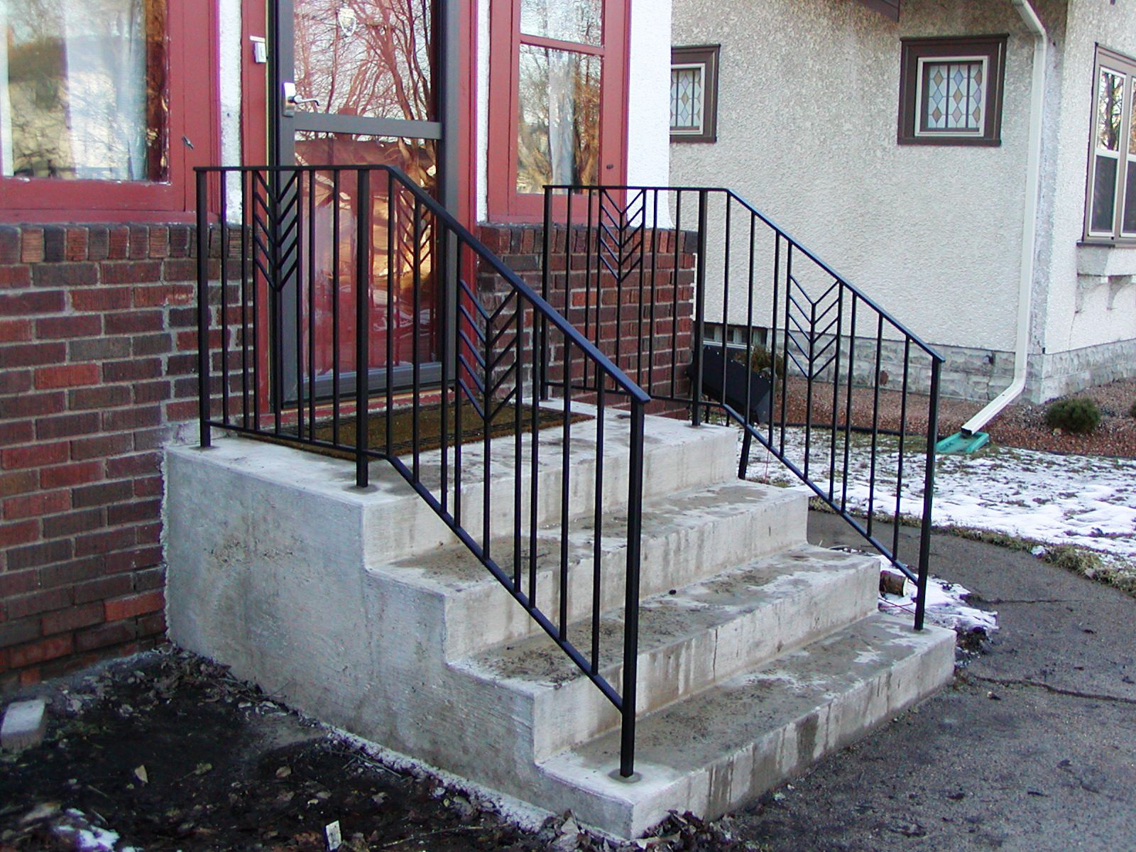 Concrete steps leading to a red door with black iron handrails. Snow on the ground.
