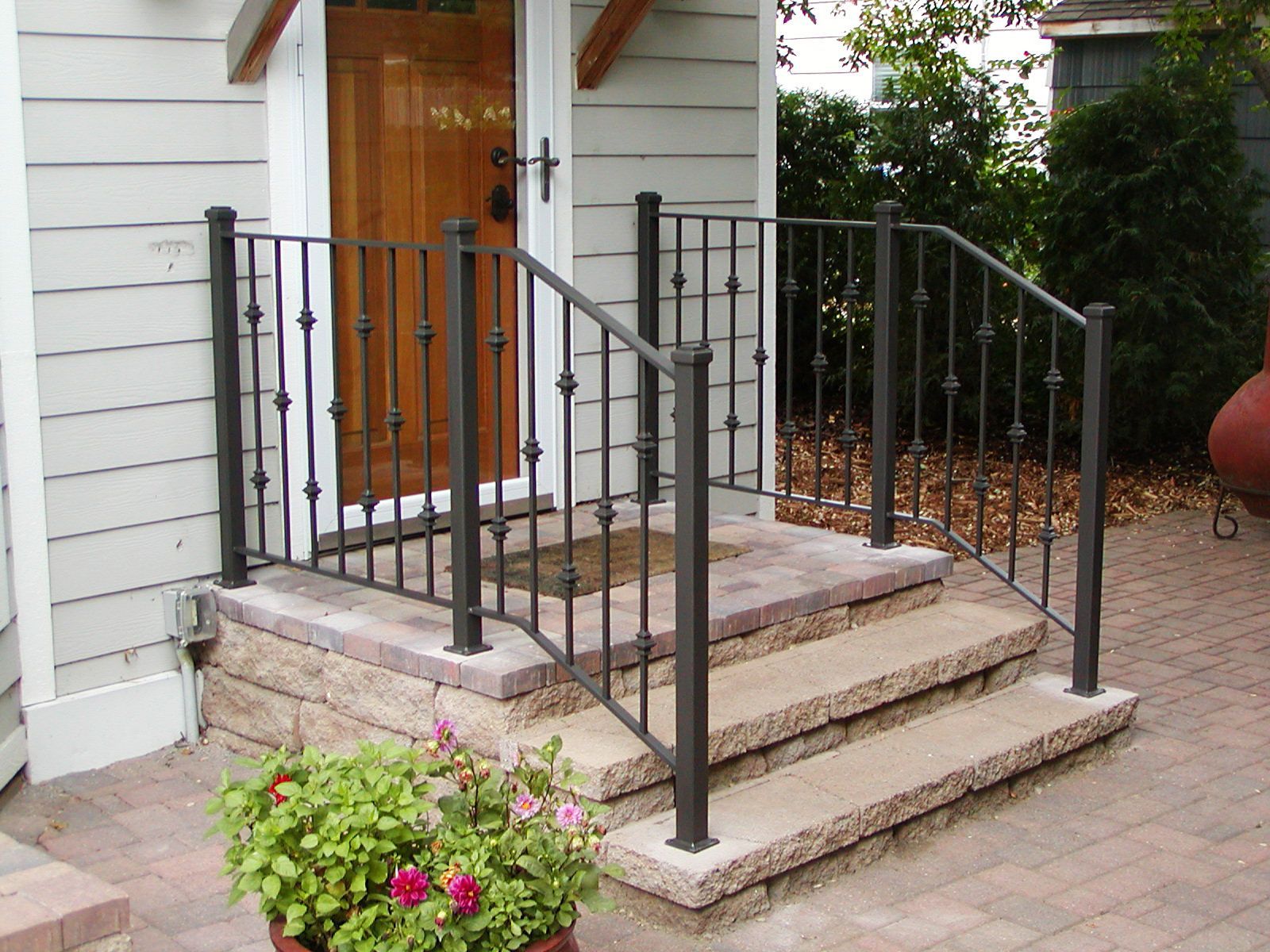 Iron railing and steps leading to a home's front door; brown door, light gray siding, flower pot.