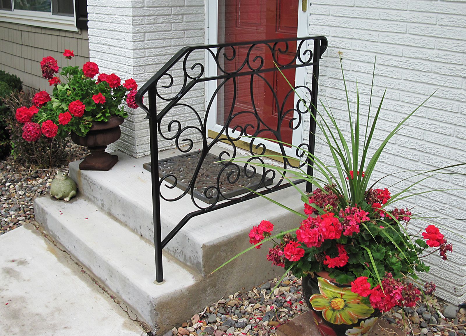 Concrete steps leading to a red door, flanked by potted red flowers and a decorative black wrought-iron handrail.