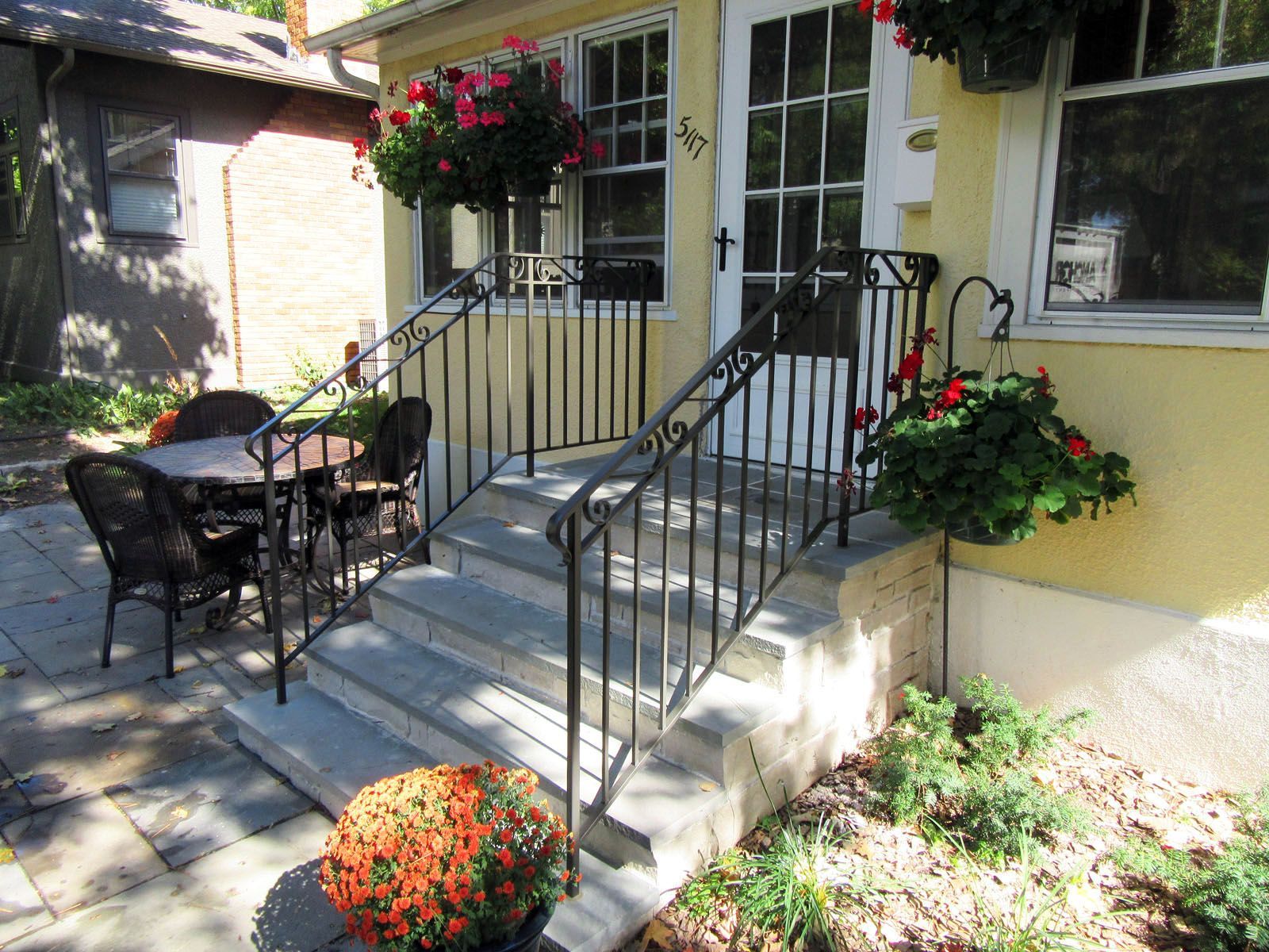 A light-colored building with steps, a black railing, and hanging red flowers, with outdoor seating on the left.
