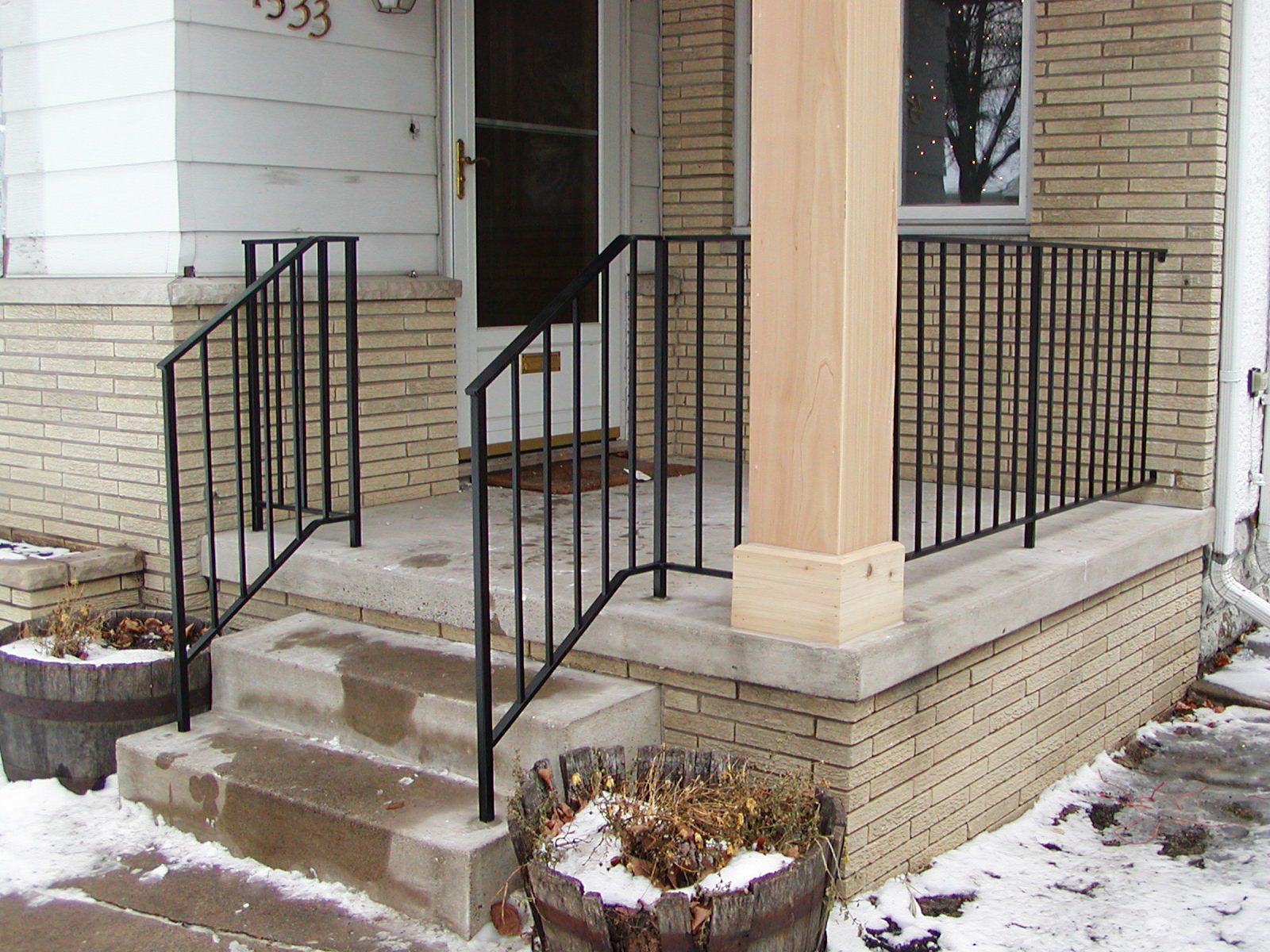 Black metal railing on a concrete porch with steps, a wooden column, and a brick wall. Snow present.
