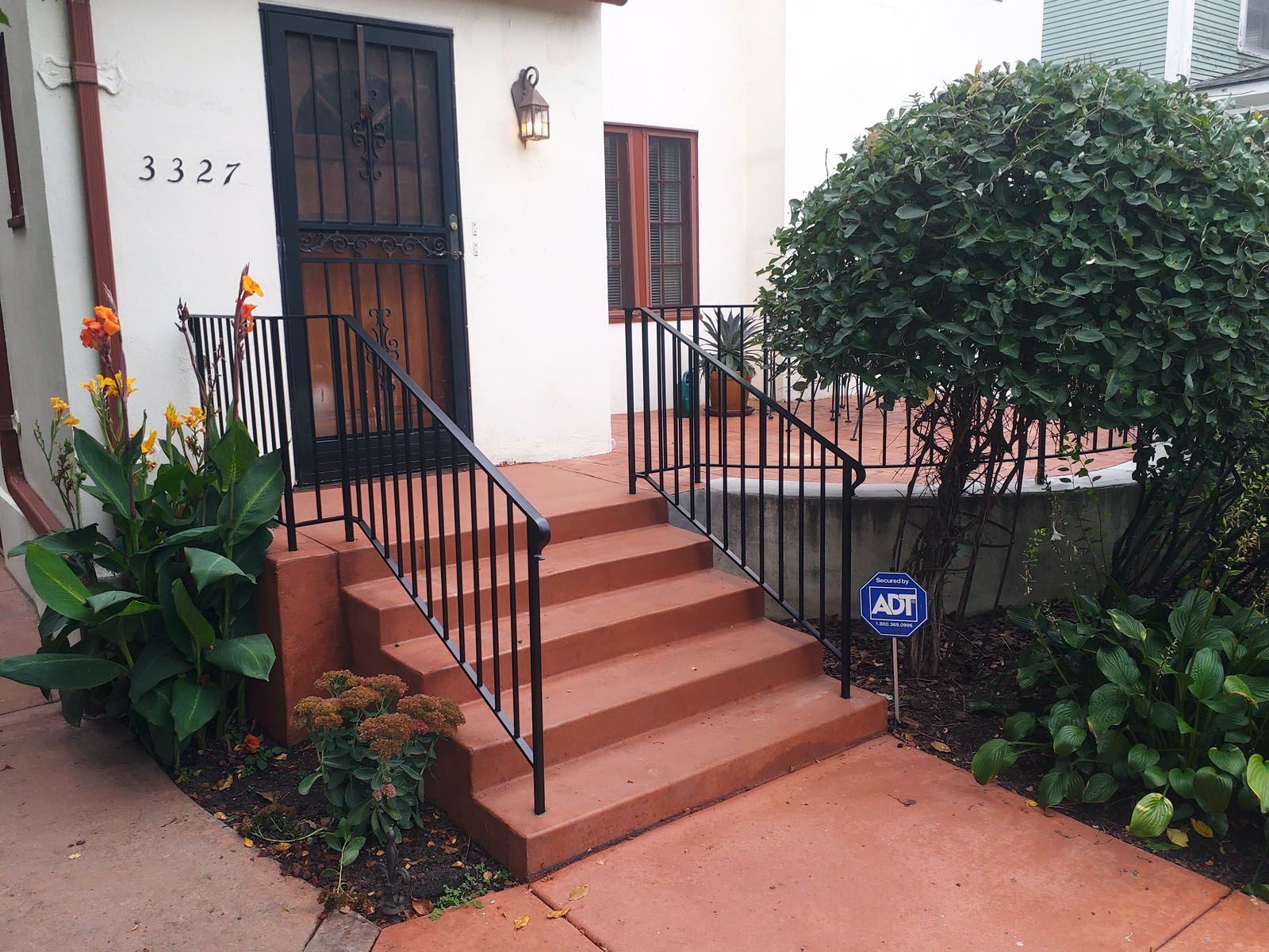 Entrance with steps and black metal railings leading to a door with a security gate; a bush and flowers nearby.