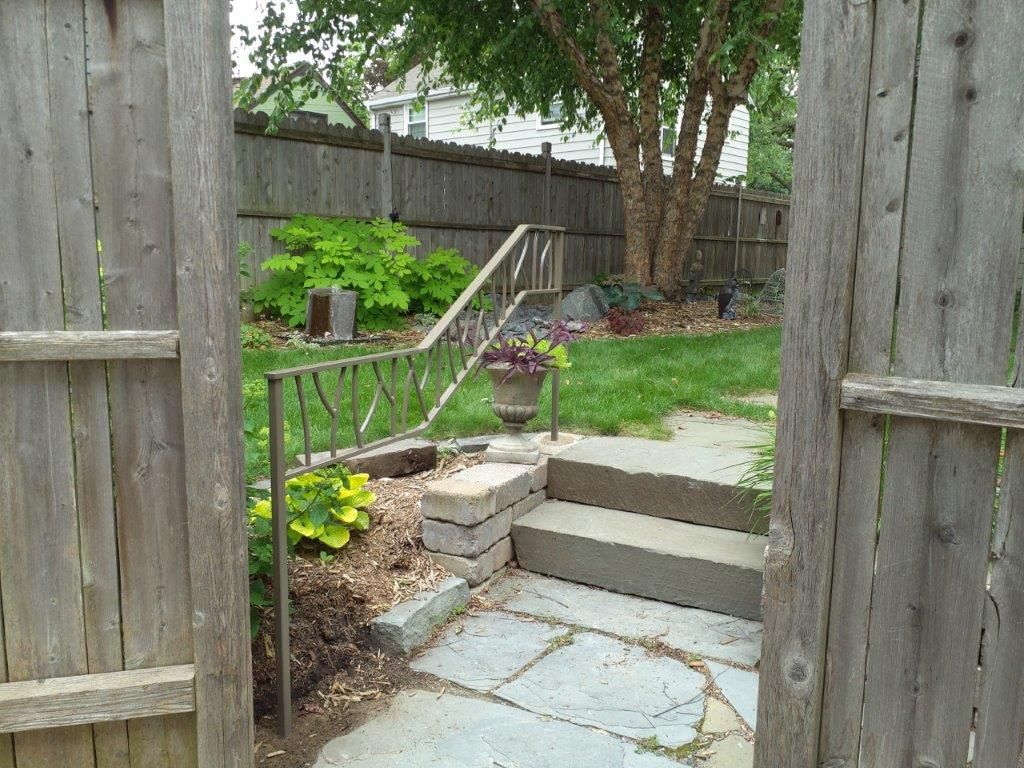 View through open wooden gate to backyard with steps, grass, and trees.
