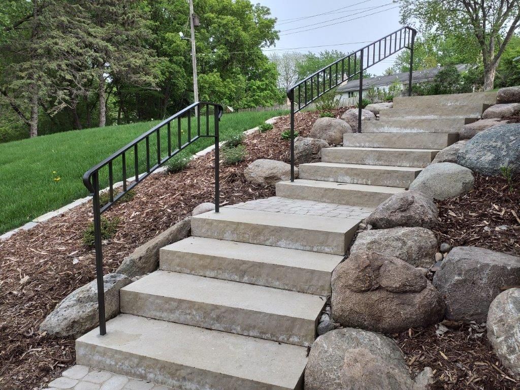 Concrete stairs with black railing ascending a hillside, surrounded by rocks and landscaping.