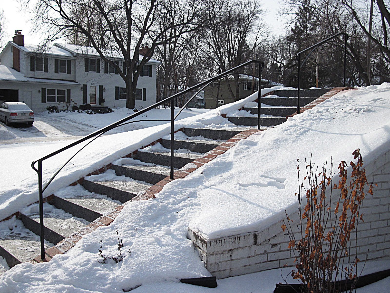 Snow-covered outdoor staircase with black handrails leading up to a house on a winter day.