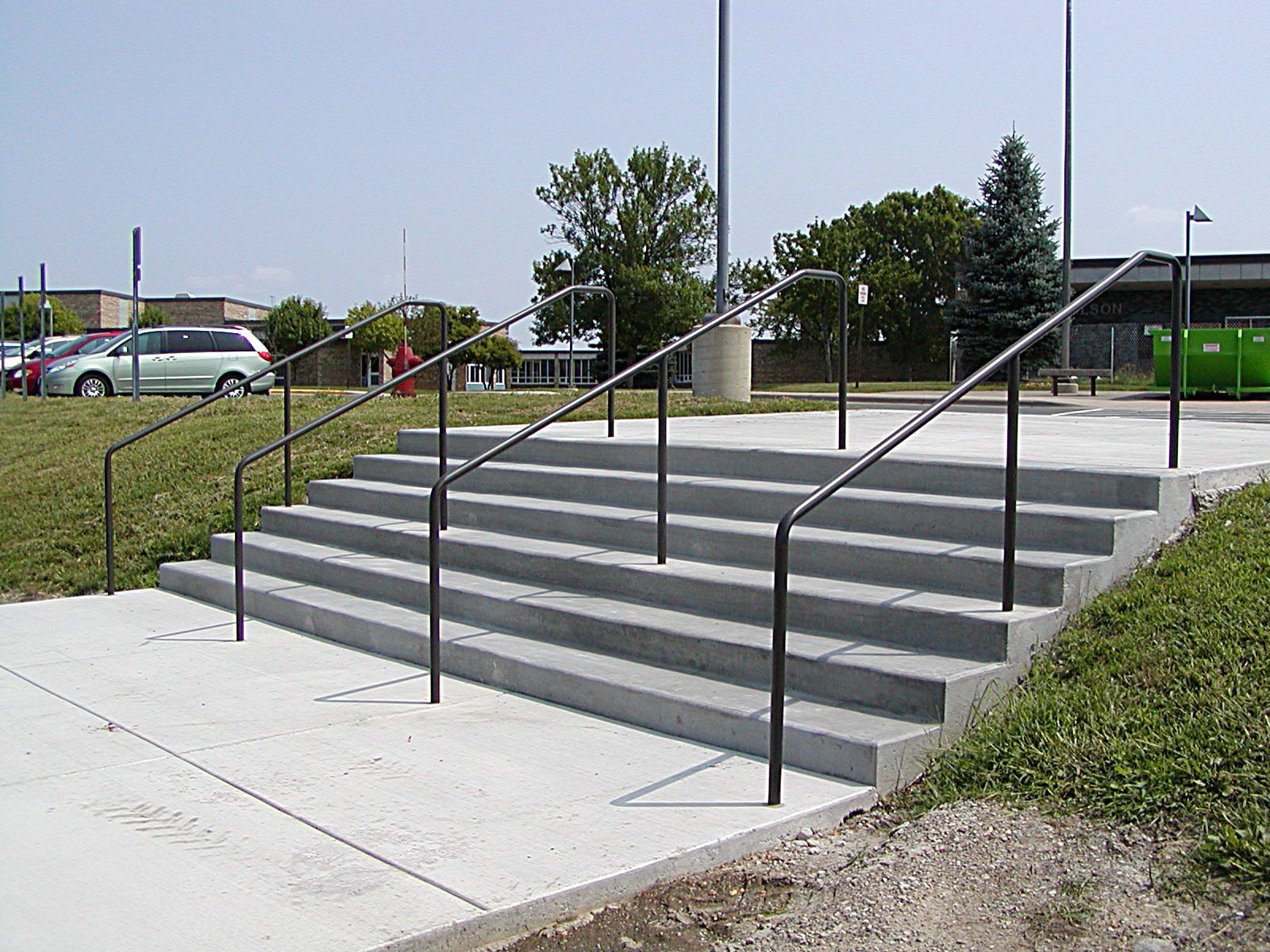 Concrete staircase with black handrails leading up to a platform. Exterior, sunny day.