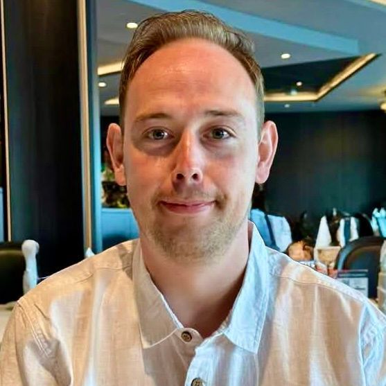 Man in a white button-down shirt smiles, seated indoors, dining.