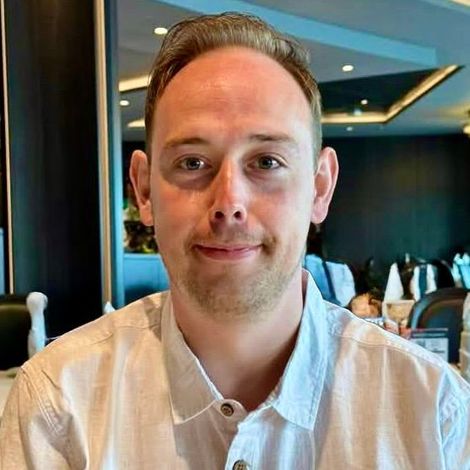 Man in a white button-down shirt smiles, seated indoors, dining.