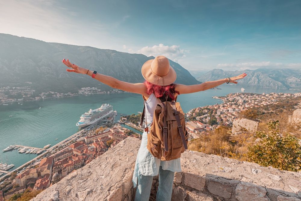 Woman with arms outstretched atop a stone wall, overlooking a bay with a city, mountains, and a cruise ship.