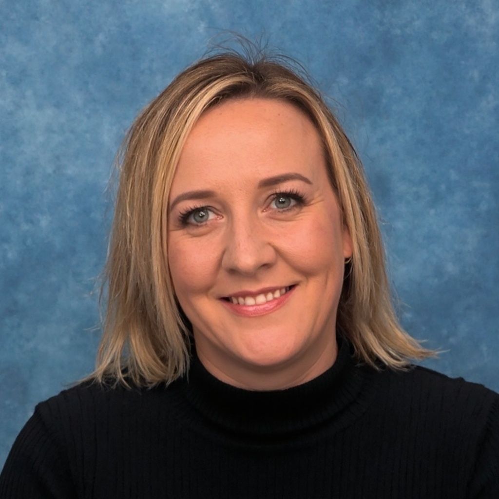 Blonde woman smiles at the camera, wearing a black turtleneck, in front of a blue background.