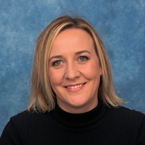 Blonde woman smiles at the camera, wearing a black turtleneck, in front of a blue background.