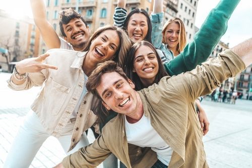 Group of friends smiling and taking a selfie outside in a city, arms raised, sunlit.