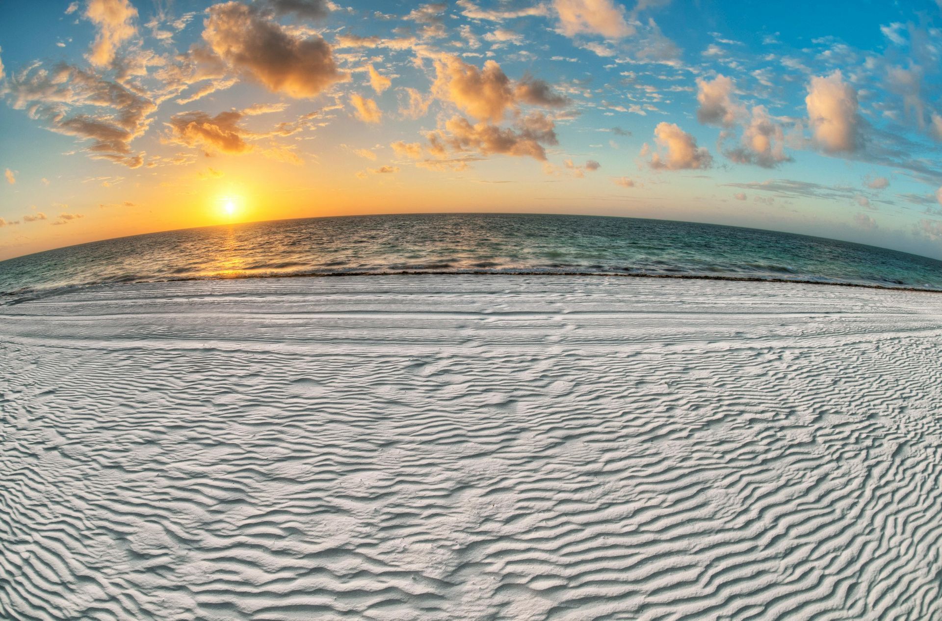 Sunset over ocean with rippled white sand beach. Blue sky with clouds.