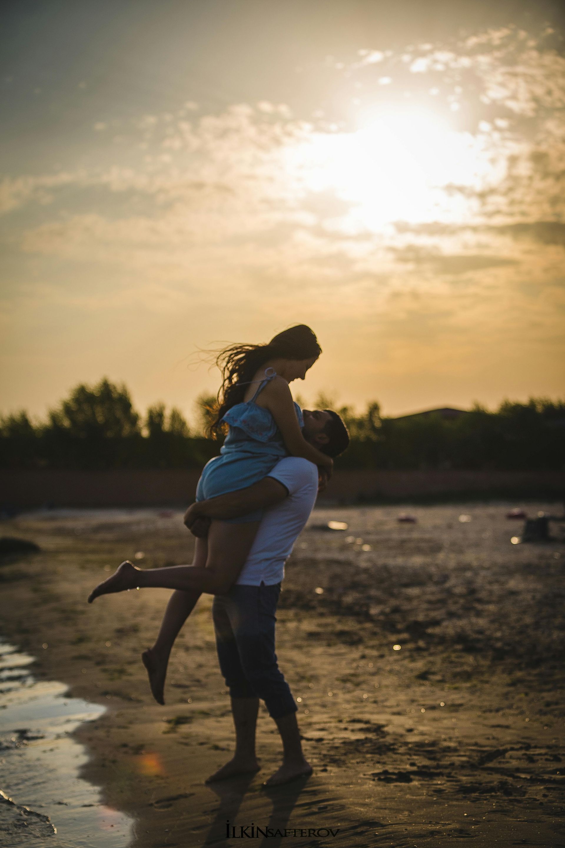 Man lifts woman on a beach at sunset. Sky is yellow, both are silhouetted, water nearby.