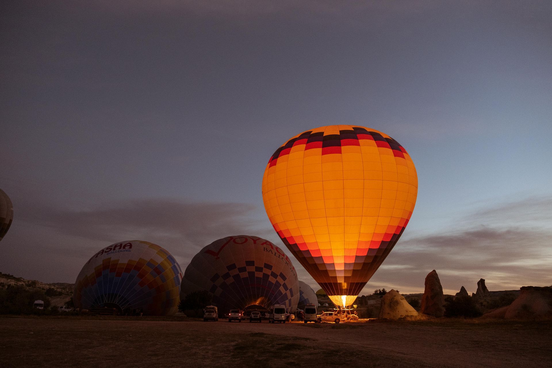 Hot air balloon glows brightly at dusk, other balloons and landscape in background.