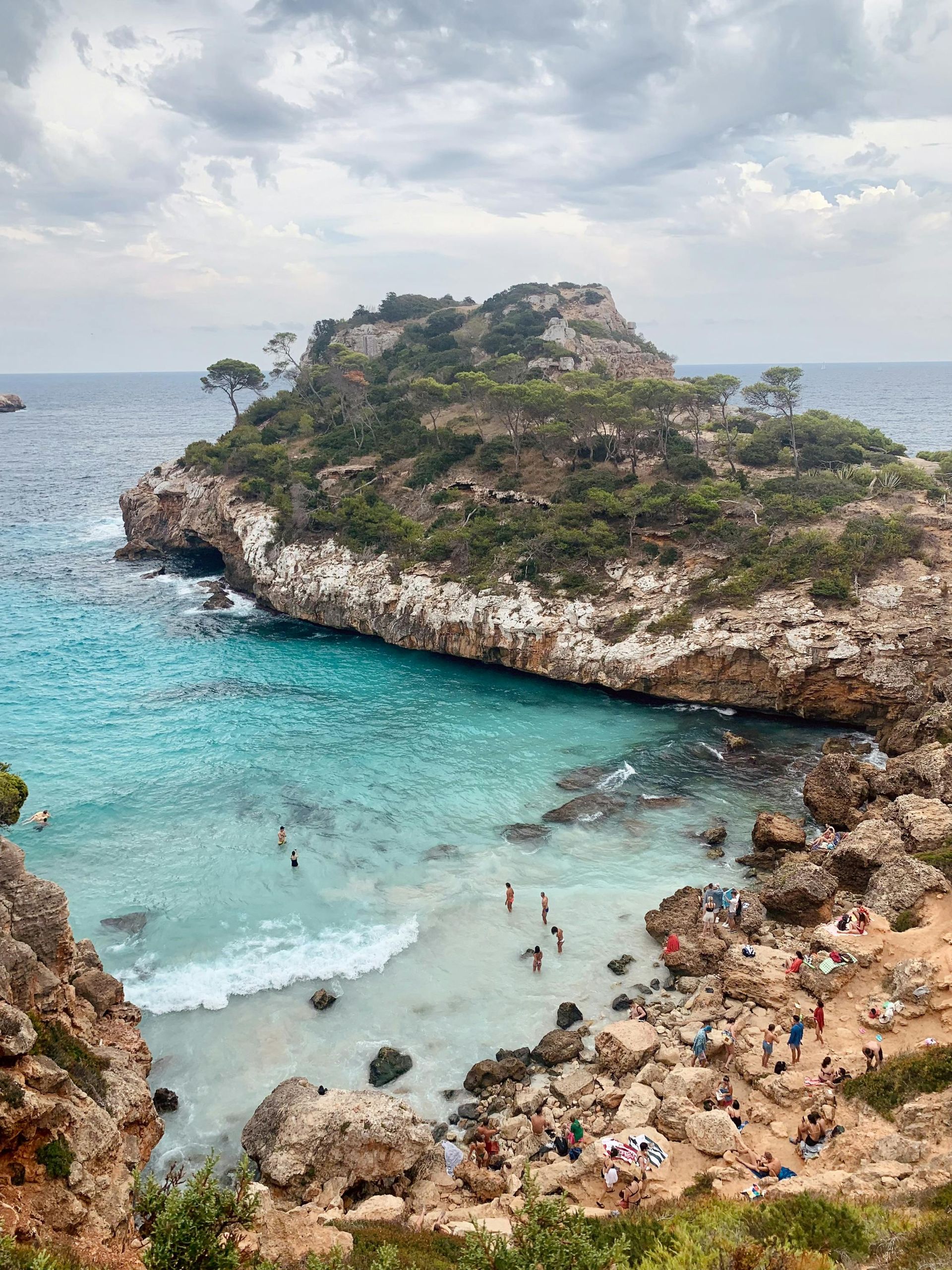 Coastal view of a small island with turquoise water. People swim and sunbathe on the rocky shore.