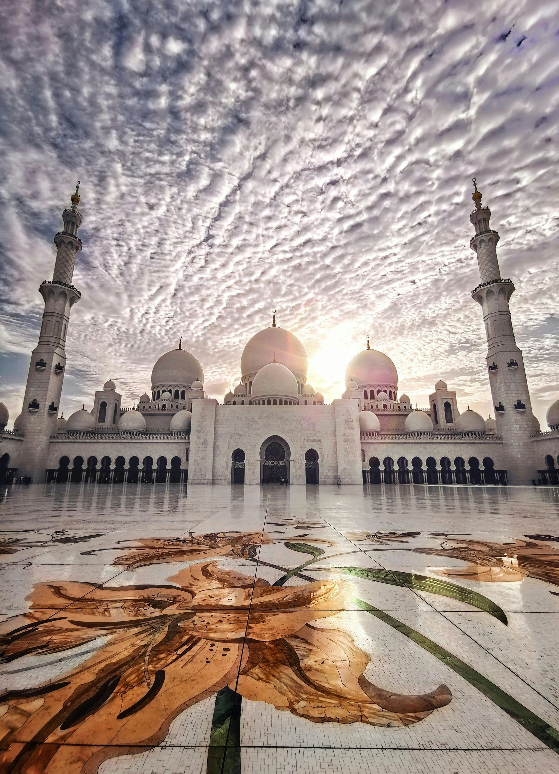 White mosque with domes and minarets, reflected on wet floor with floral design, under a cloudy sky.