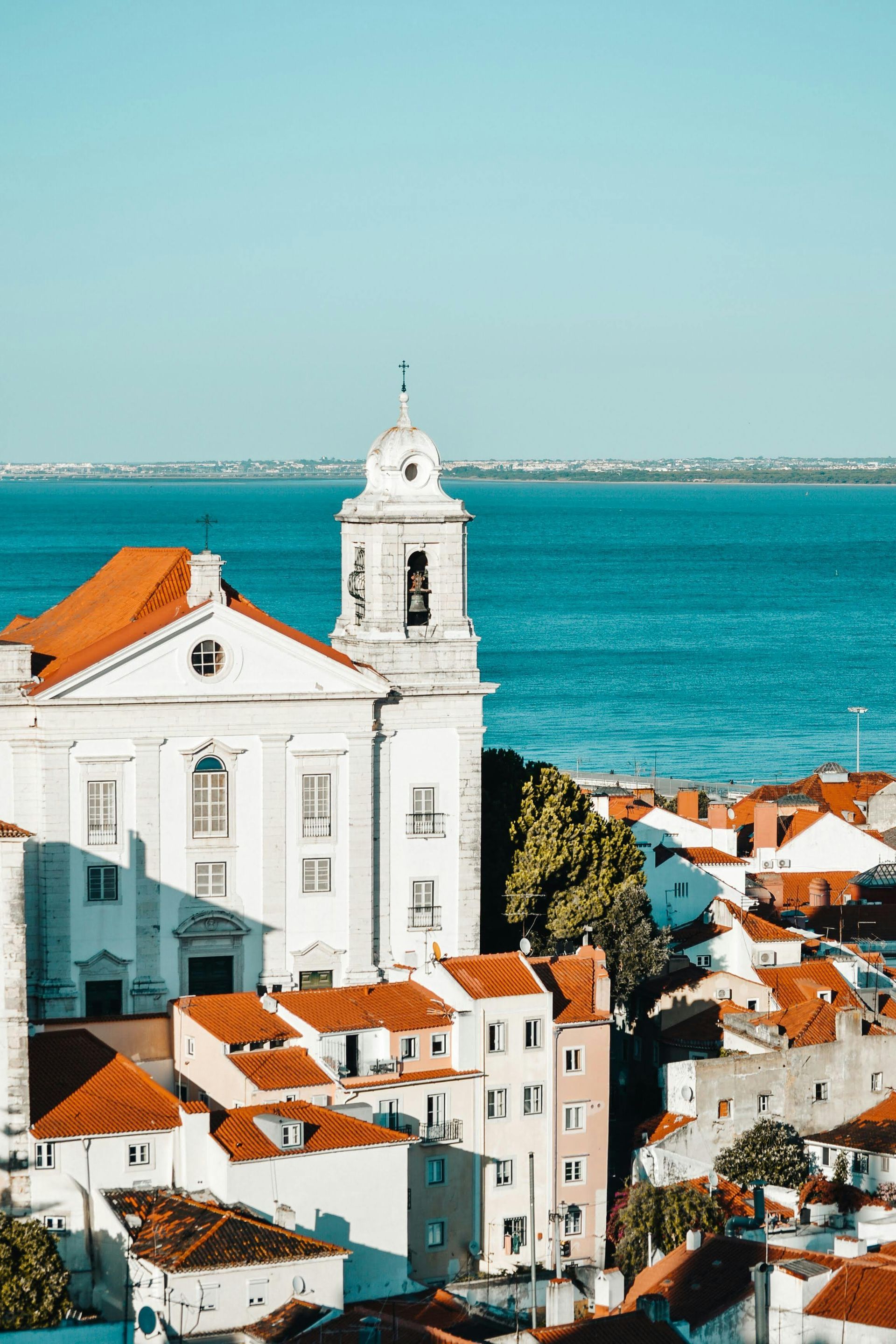 White church with orange tile roof, overlooking a city and blue water under a clear sky.