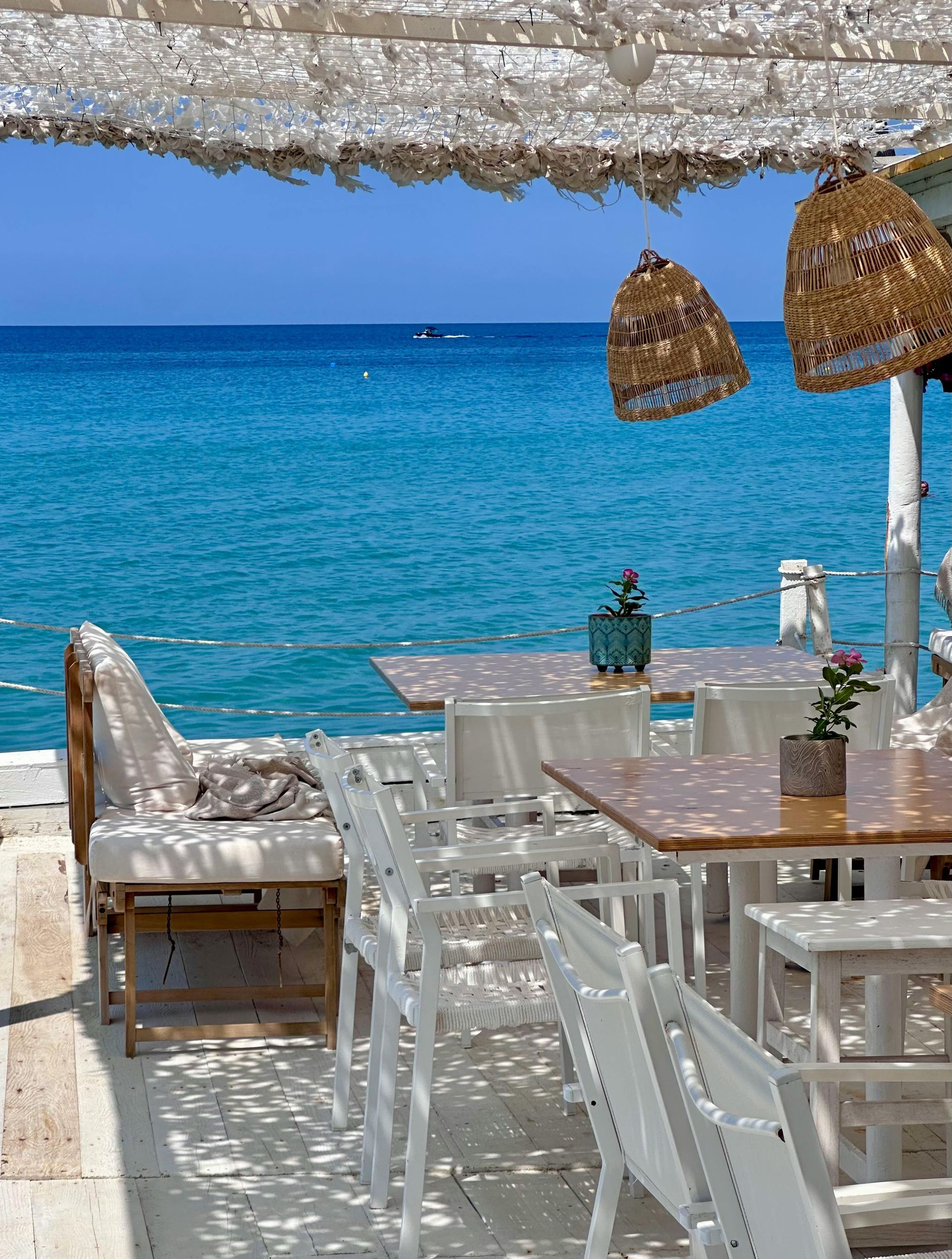 Restaurant patio by the blue sea with white furniture, woven decor, and partial shade.