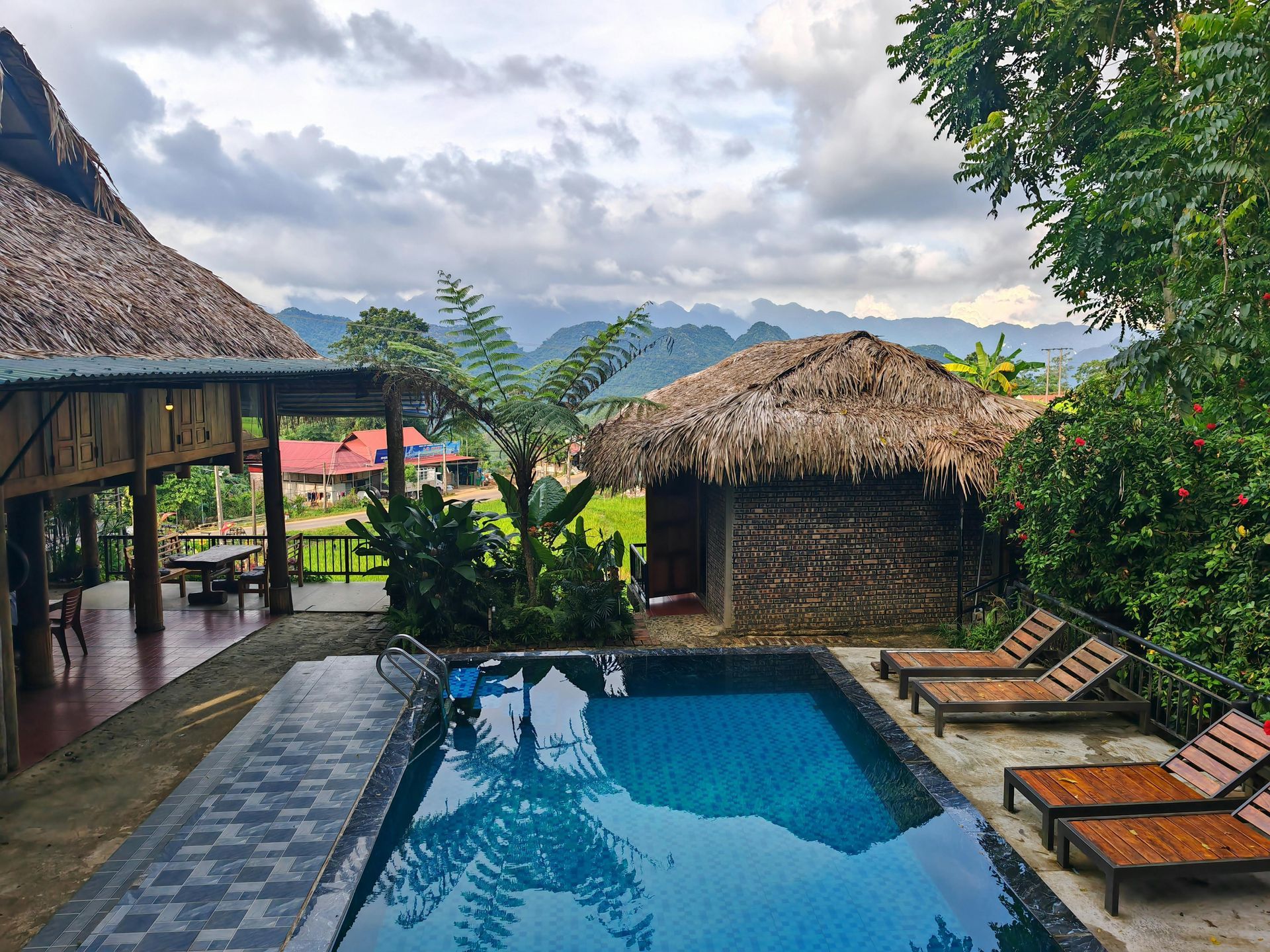 Swimming pool at a rustic resort with thatched roof buildings and mountain views.