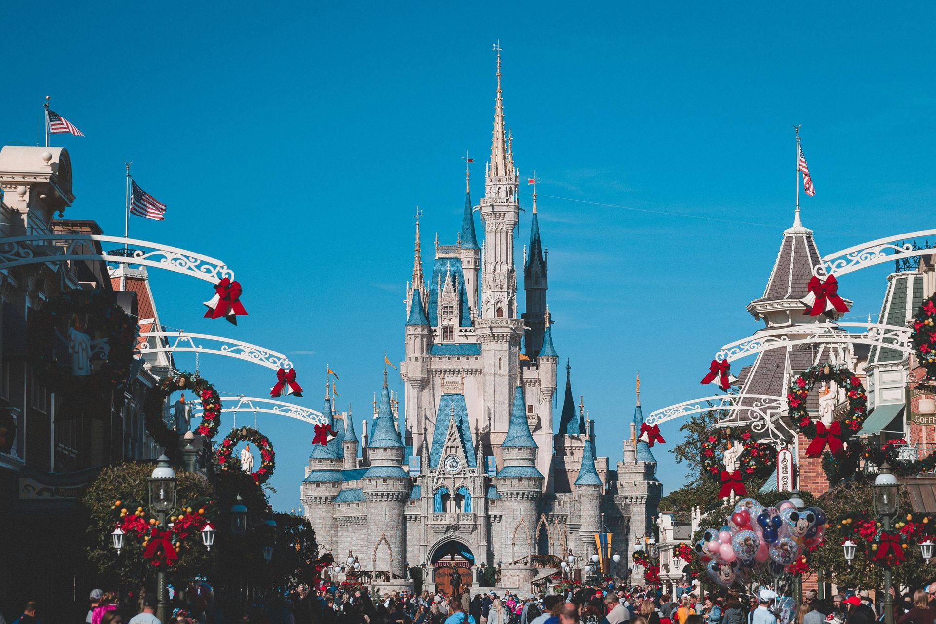 Cinderella Castle at Magic Kingdom, flanked by decorated buildings and a crowd under a clear blue sky.