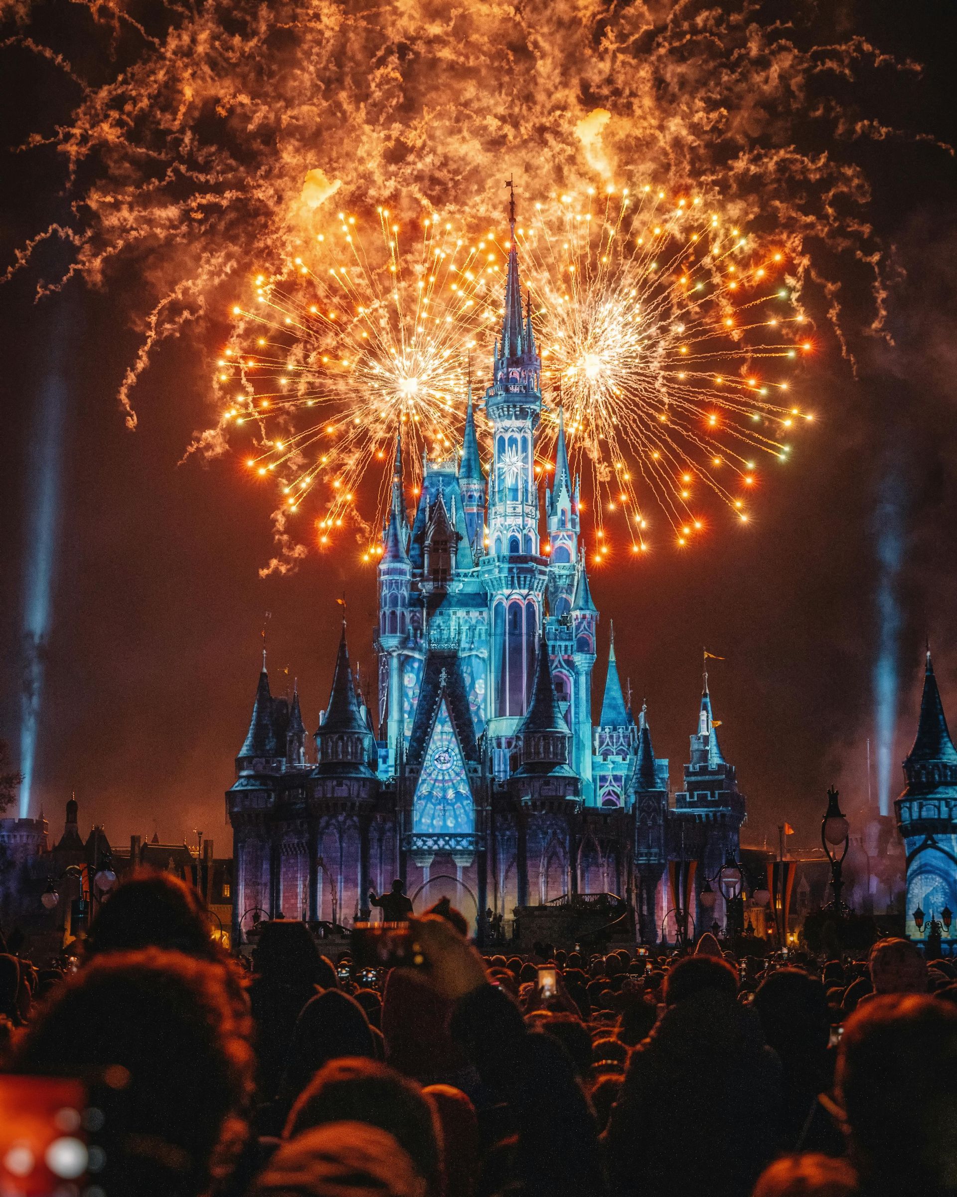 Cinderella Castle lit up with blue lights and surrounded by bright orange fireworks during a nighttime show in a theme park.