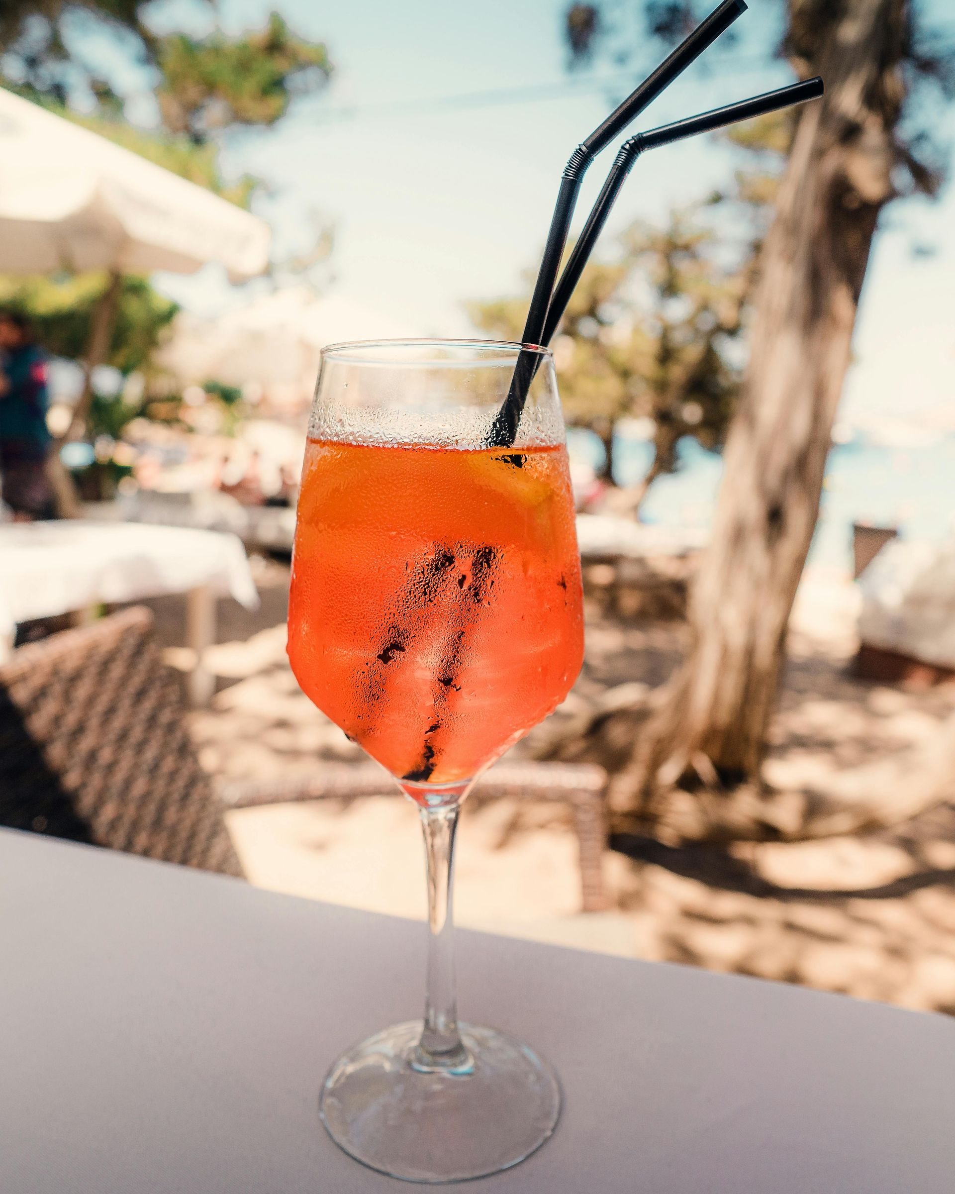 Orange cocktail in a stemmed glass with black straws on a table outdoors; beach and trees in the background.