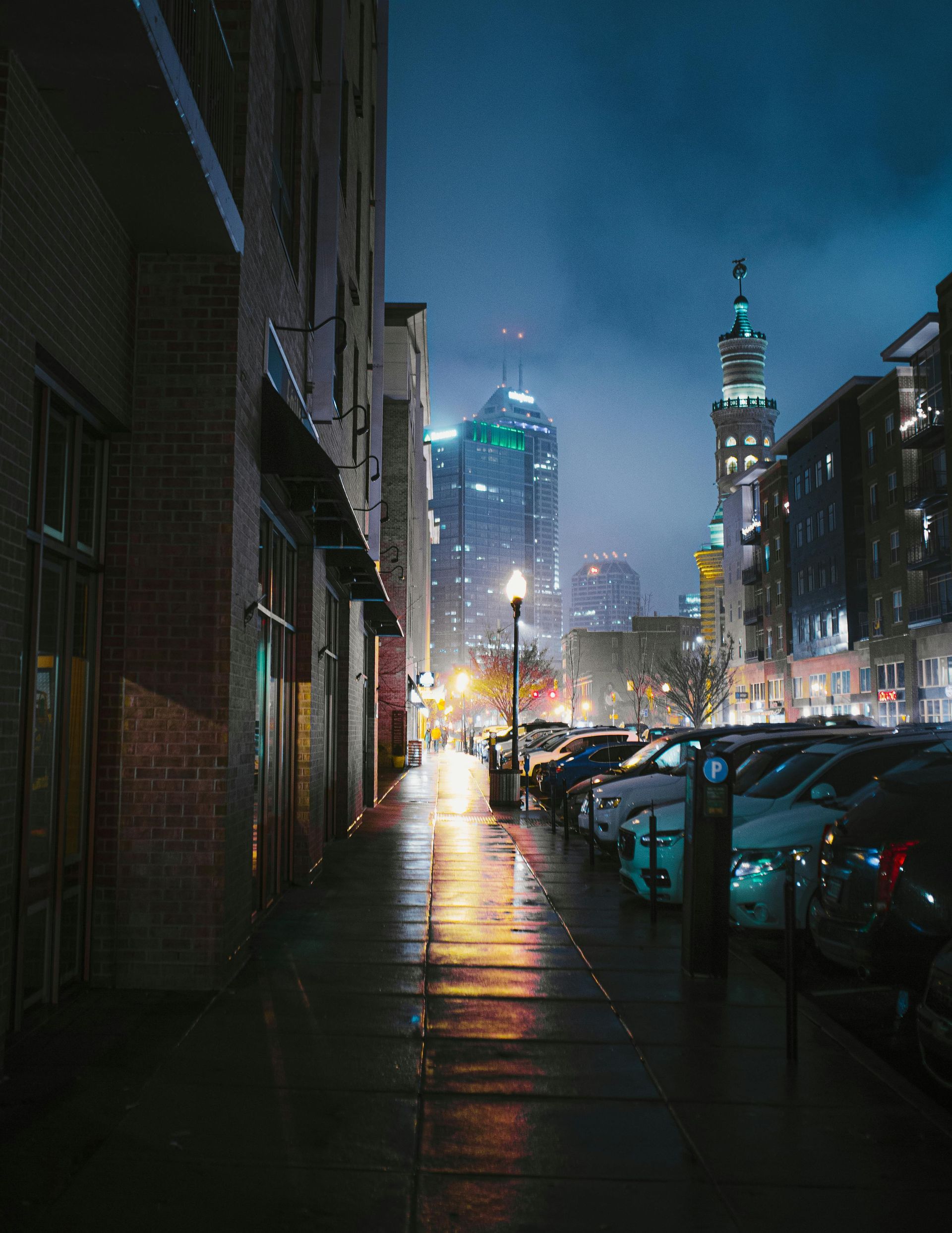 Wet city street at night, reflecting lights. Buildings and parked cars line the sidewalk, with skyscrapers in the background.