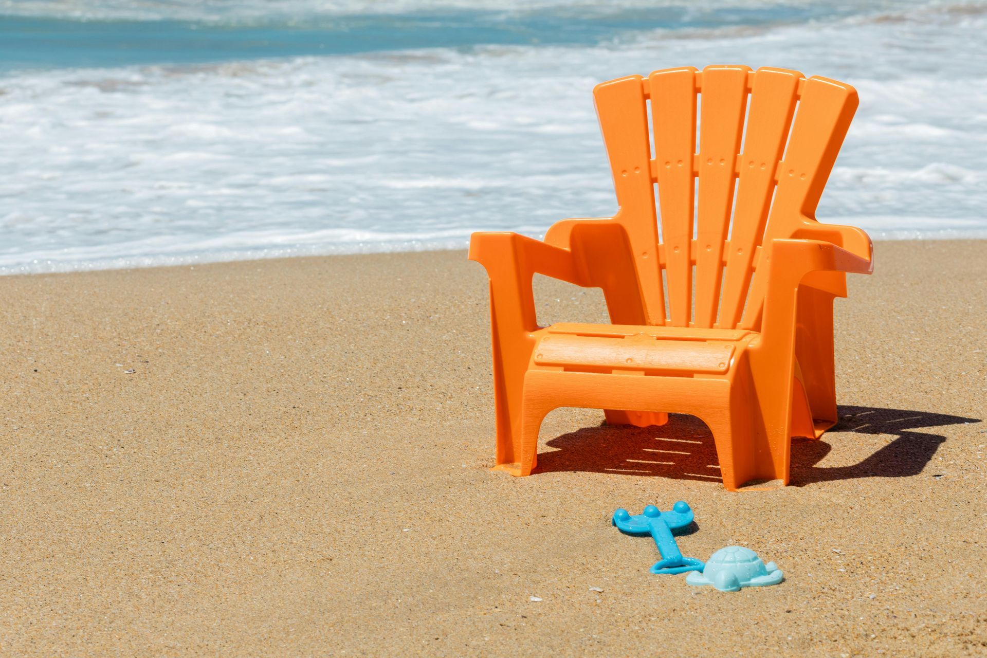 Orange Adirondack chair on a sandy beach near ocean waves, with a blue toy spade and pail.