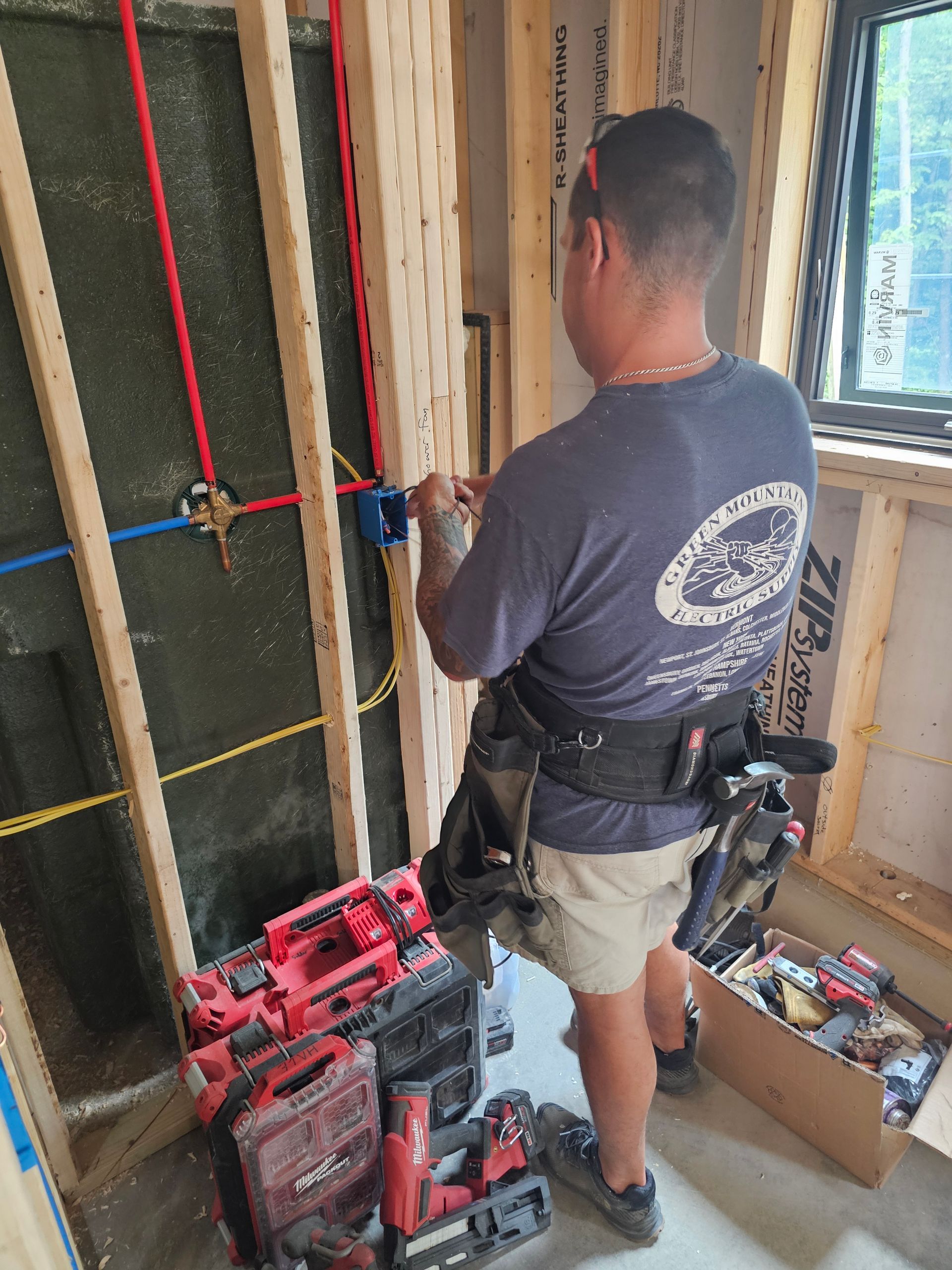 Construction worker wiring electrical box in a wood-framed wall; tools at feet.