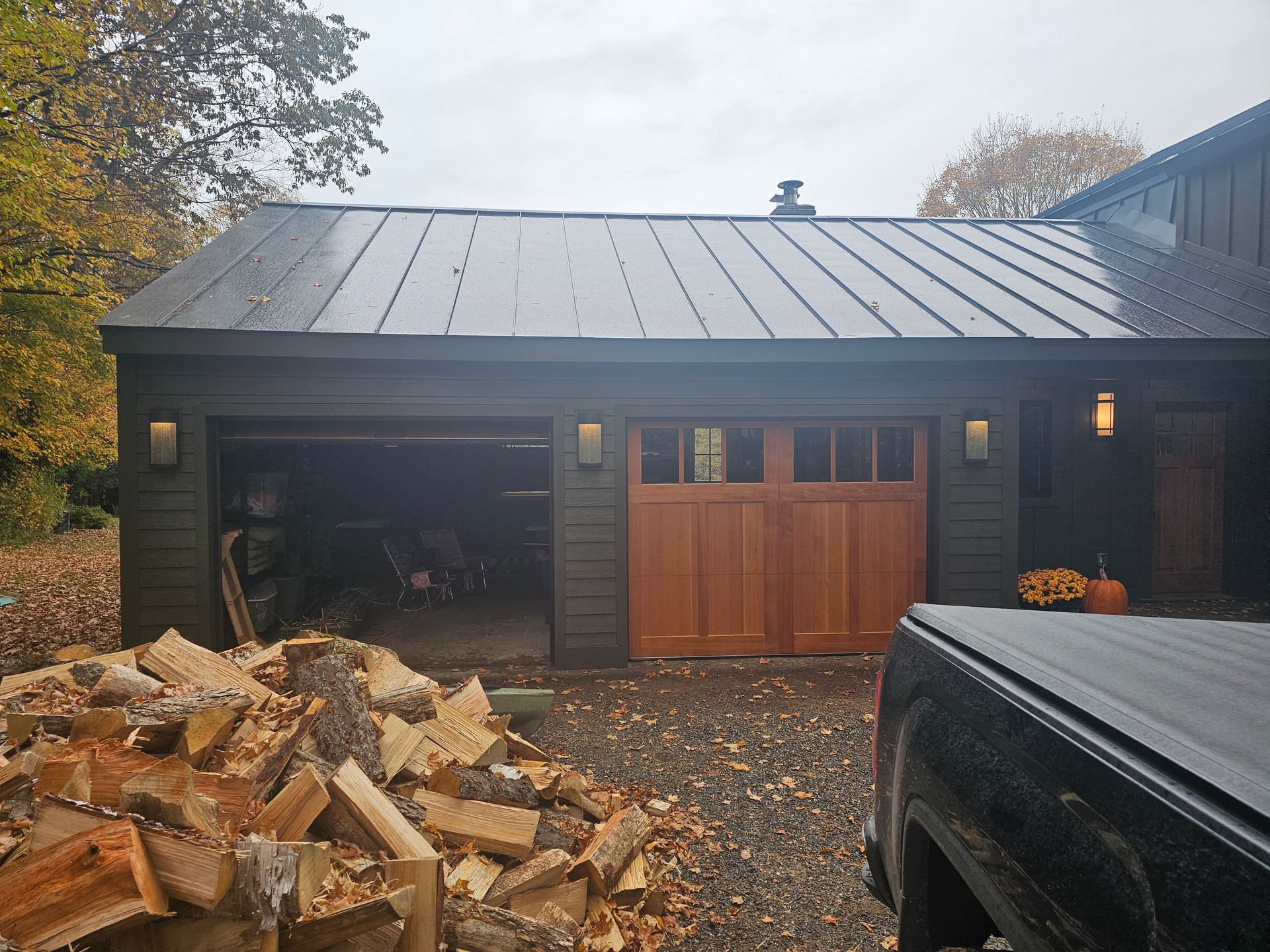 Dark green garage with brown door and metal roof; firewood and a truck in front.