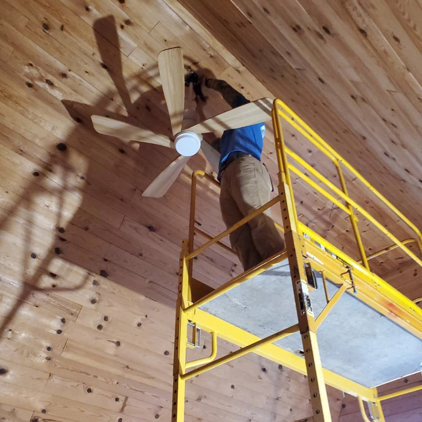 Person on a yellow lift installs a ceiling fan on a wood-paneled ceiling.