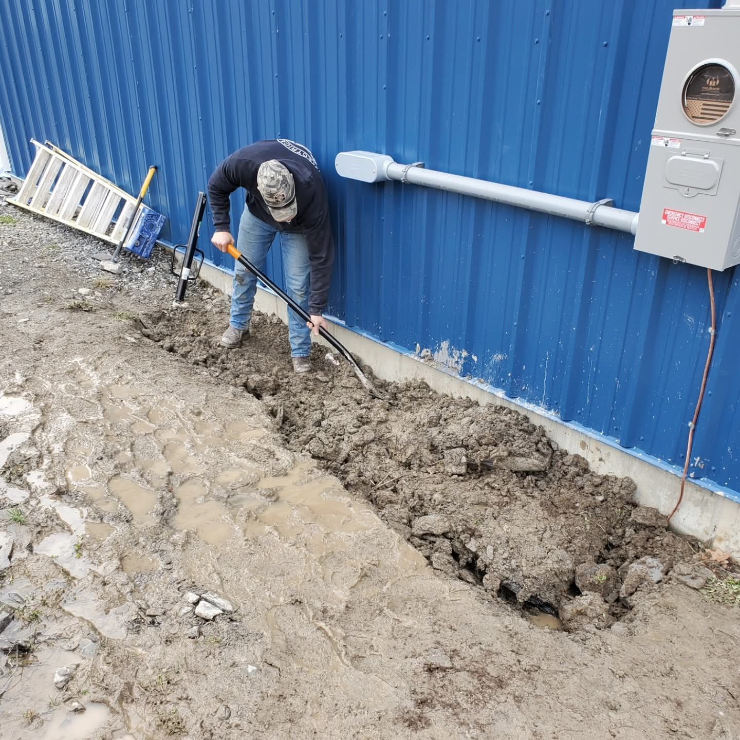 Person digging a trench next to a blue building with electrical equipment. Muddy ground.