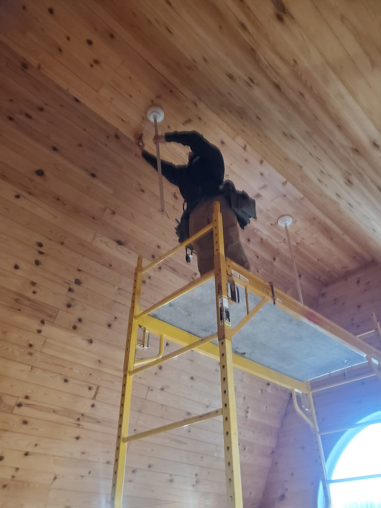 Person on a yellow scaffold installing light fixtures on a wood-paneled ceiling.