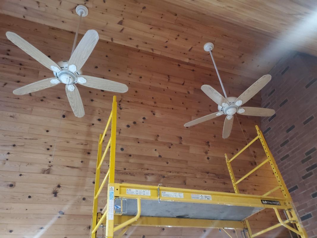 Two ceiling fans installed on a wood plank ceiling; a yellow scaffold stands beneath the fans.