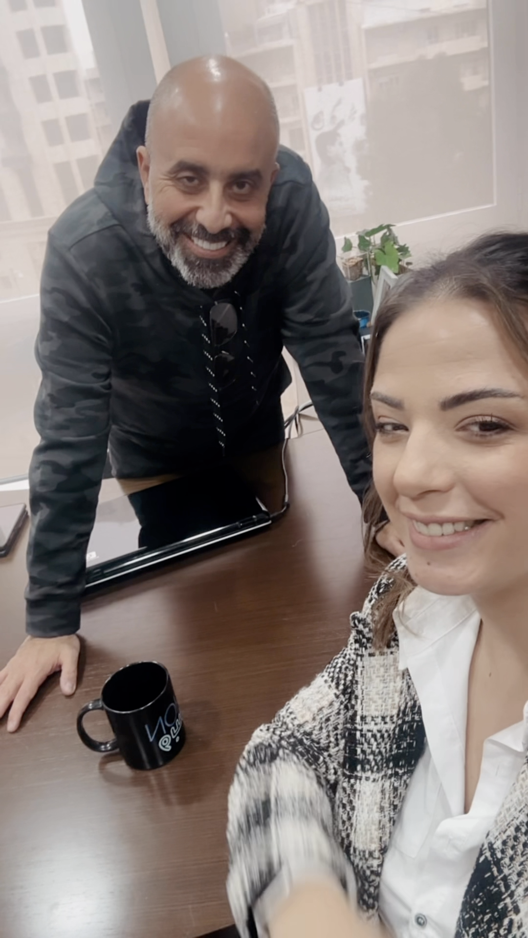 A woman takes a selfie with a smiling man. They stand by a desk in an office.