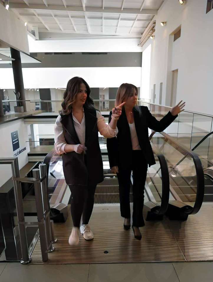 Two women in business attire walking near an escalator, one pointing to the right.