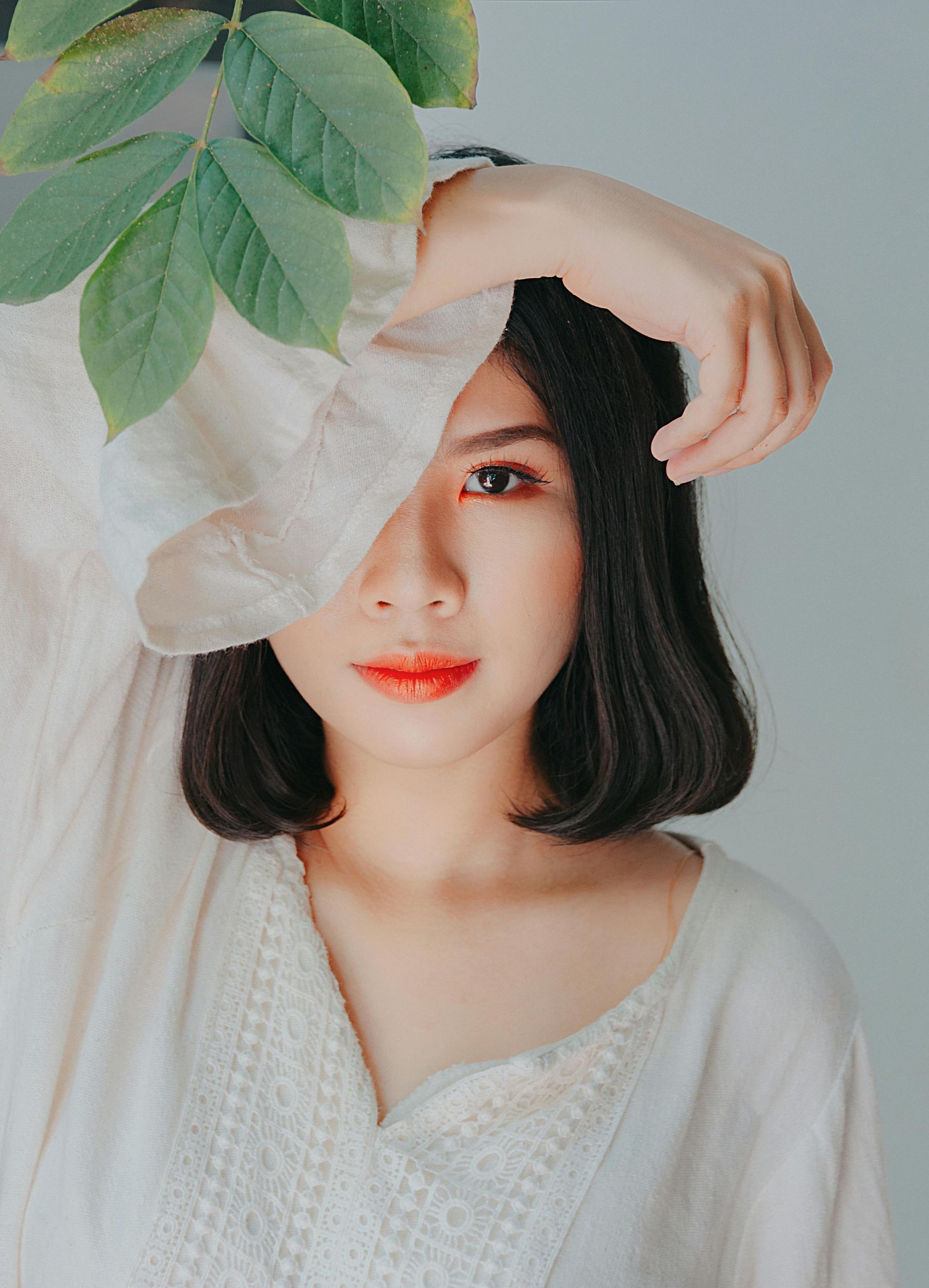 Woman with bob haircut, wearing white top, arm raised to face near green leaves.