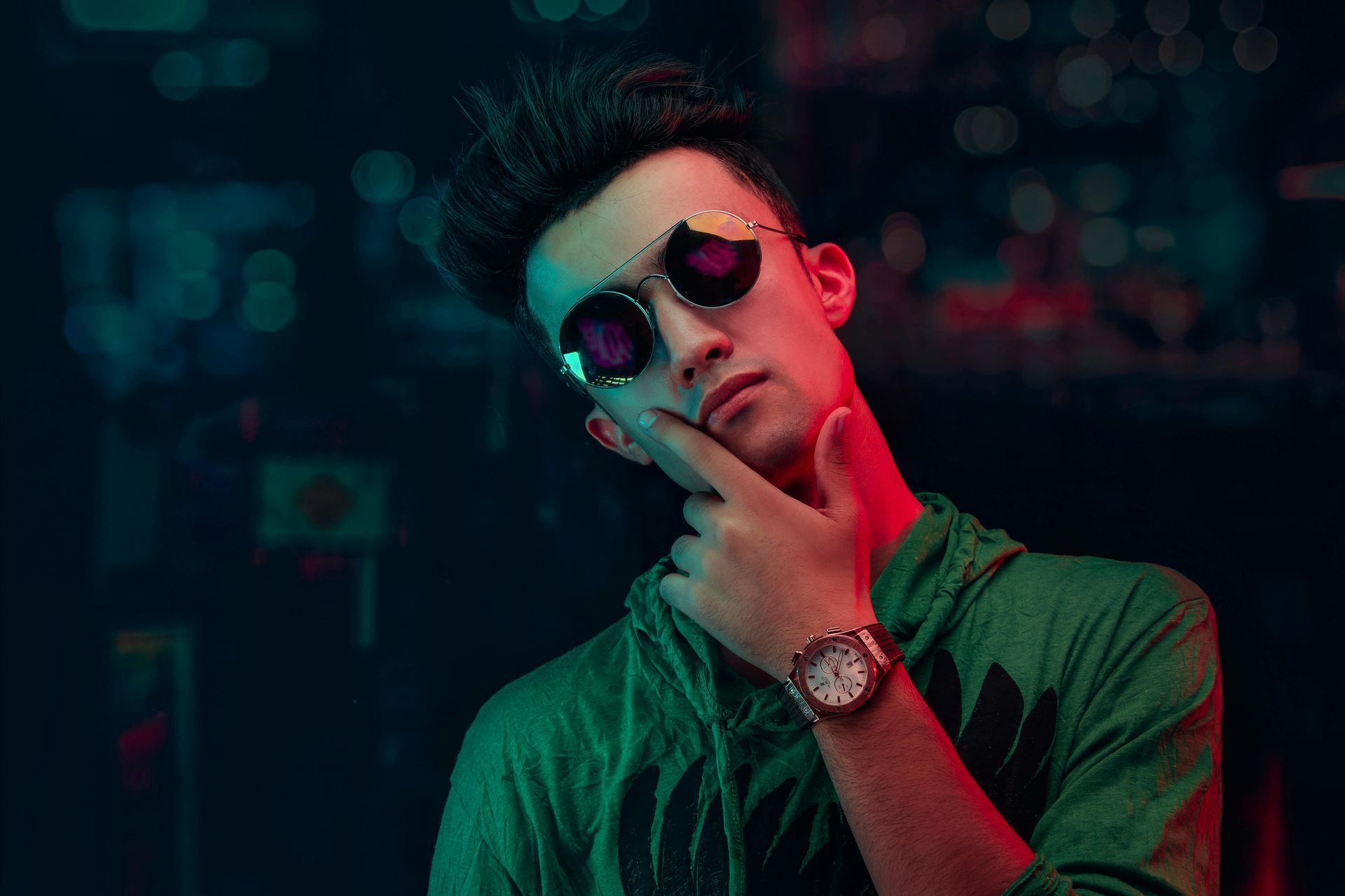 Man with sunglasses, holding chin, city background lit with red and green.