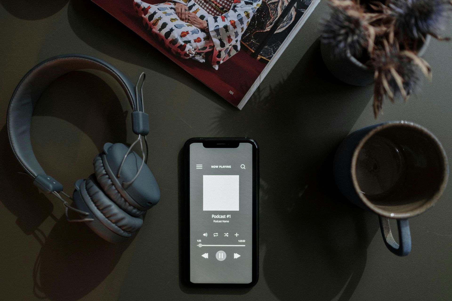 Smartphone playing music next to headphones, a mug, and a magazine on a dark surface.