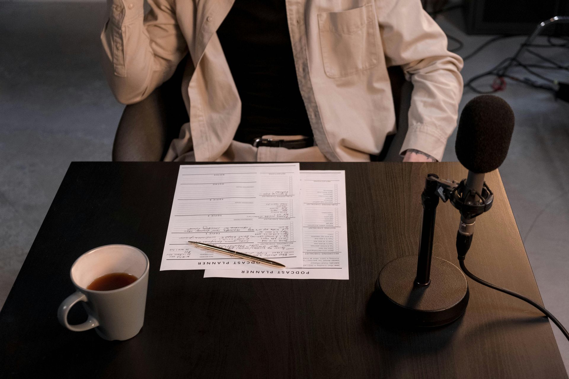 Person at desk with microphone, notes, and coffee. Beige shirt, black shirt.