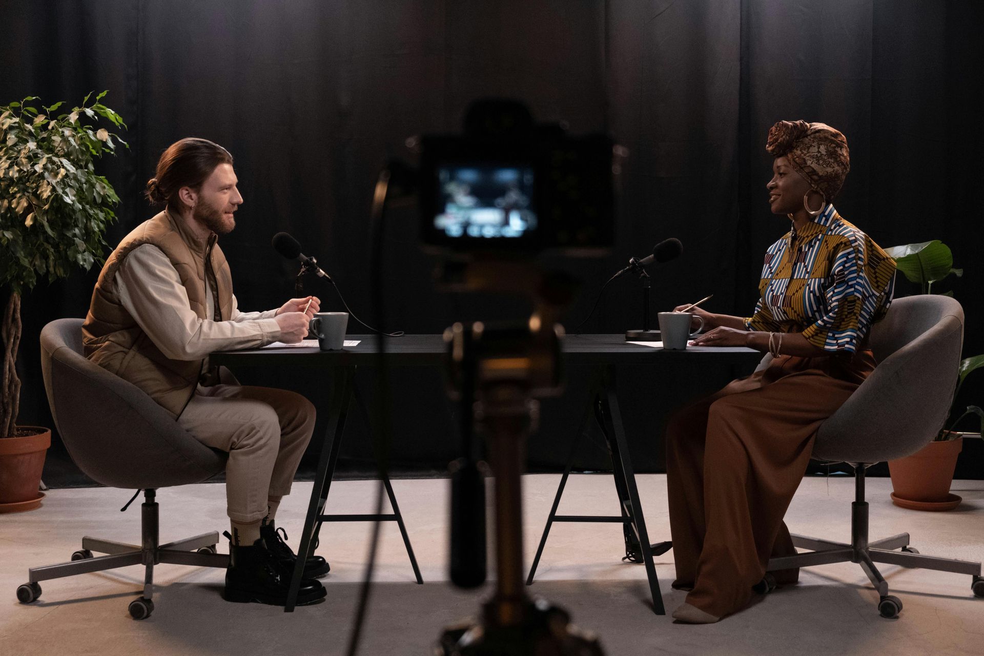 Two people sit at a table, microphones present. Dark studio background, potted plants.