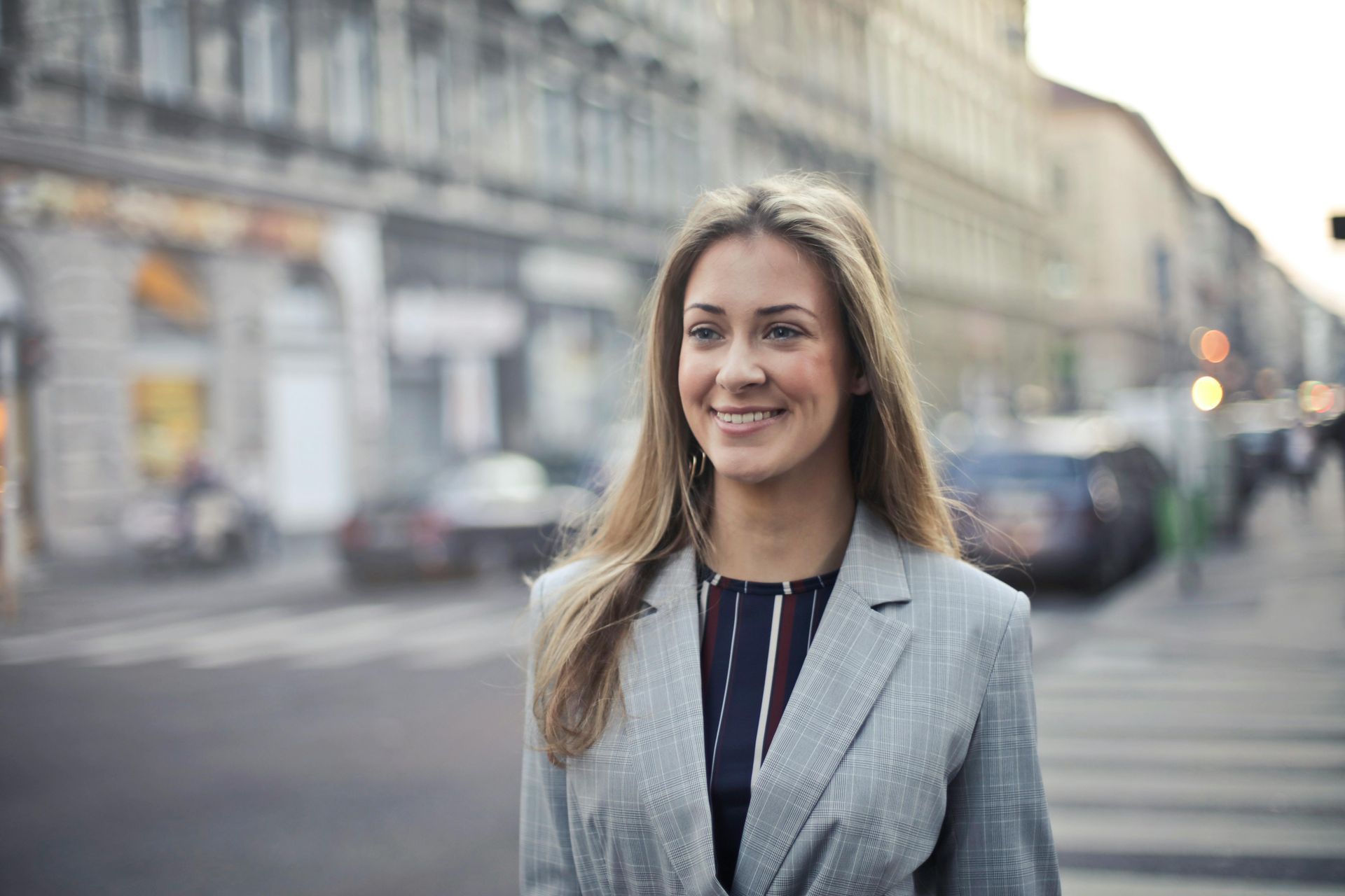 Woman in a gray blazer smiles, standing on a city street with crosswalk, cars, and buildings in the background.