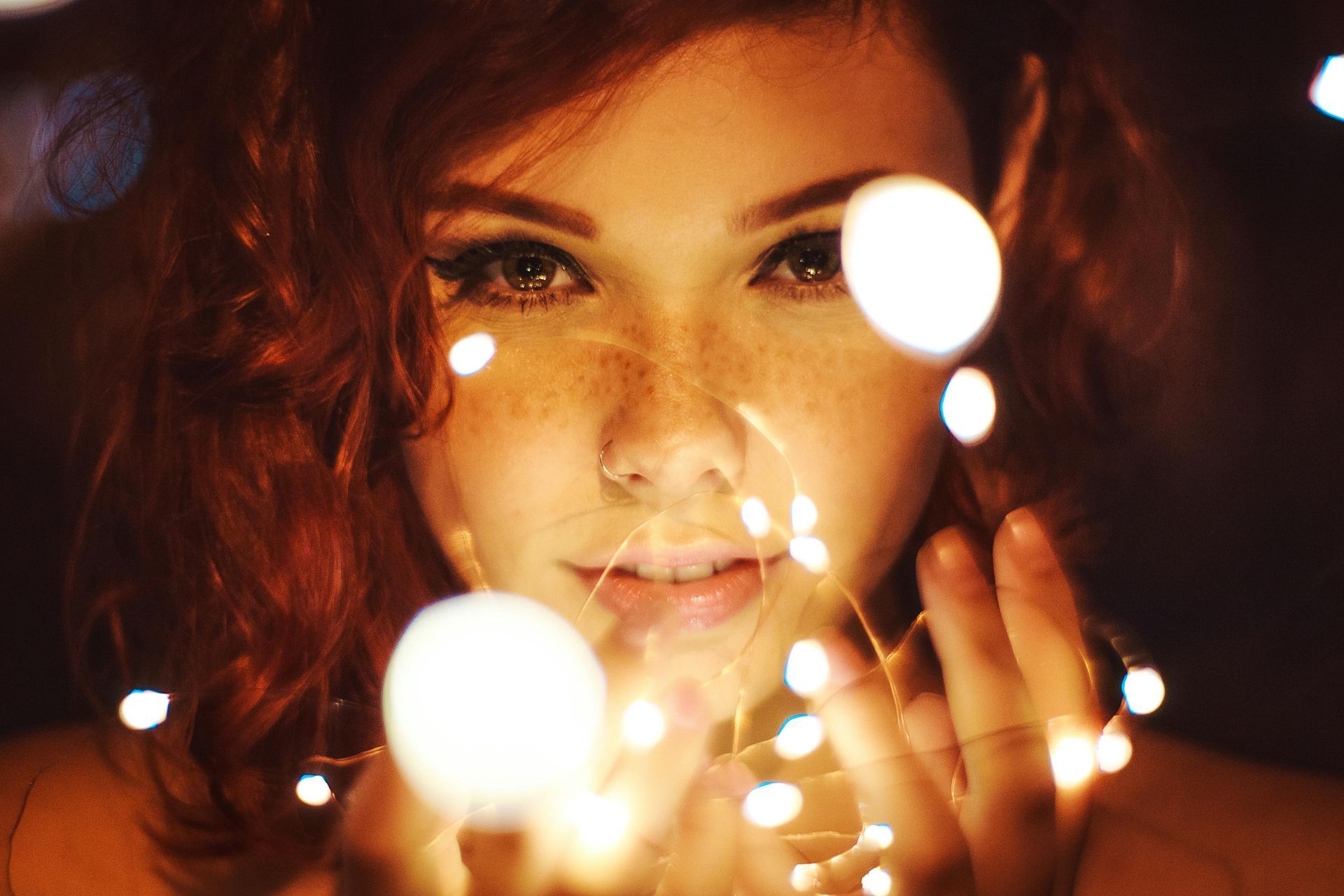 Woman with auburn hair and freckles, holding glowing fairy lights, looking toward the camera.