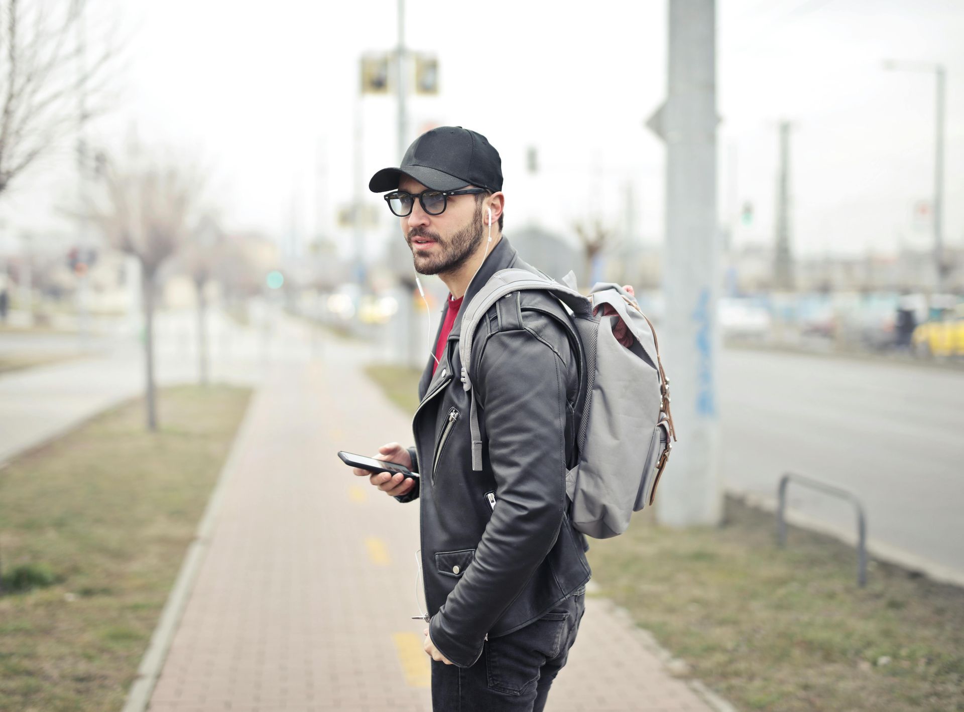Man in leather jacket and cap, holding phone, looking back on a sidewalk, backpack on.